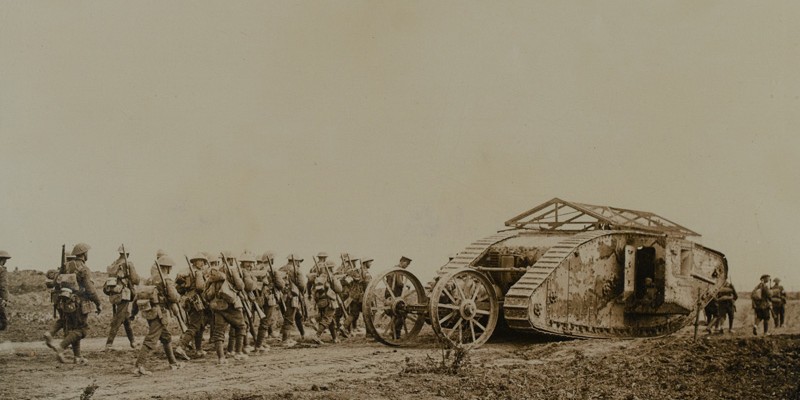 A Mark I tank with grenade screens and rear steering wheel device advances with infantry marching behind, 1916